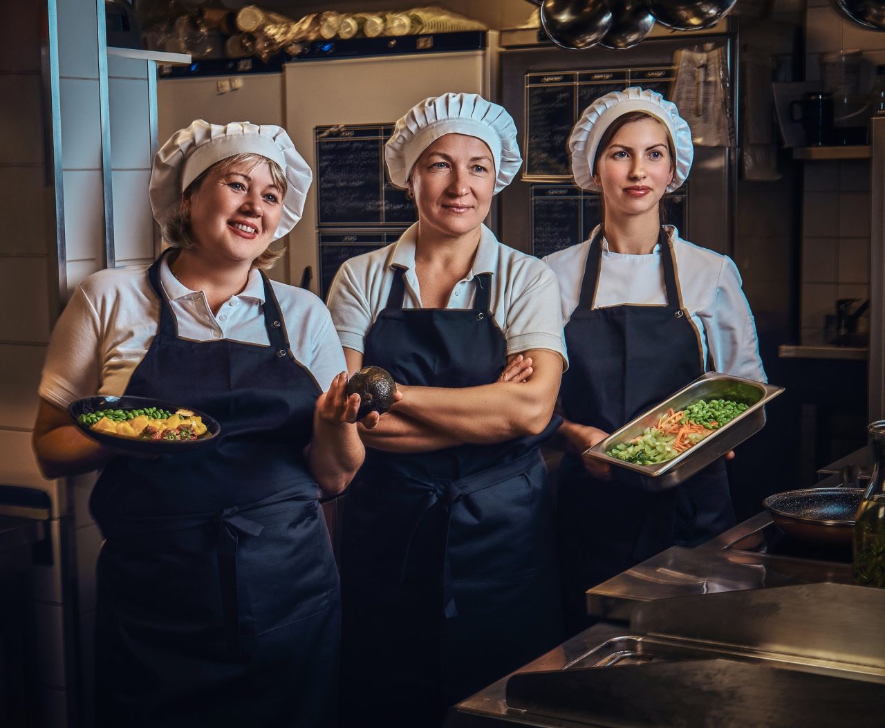 Three chefs holding fresh vegetables in a kitchen.